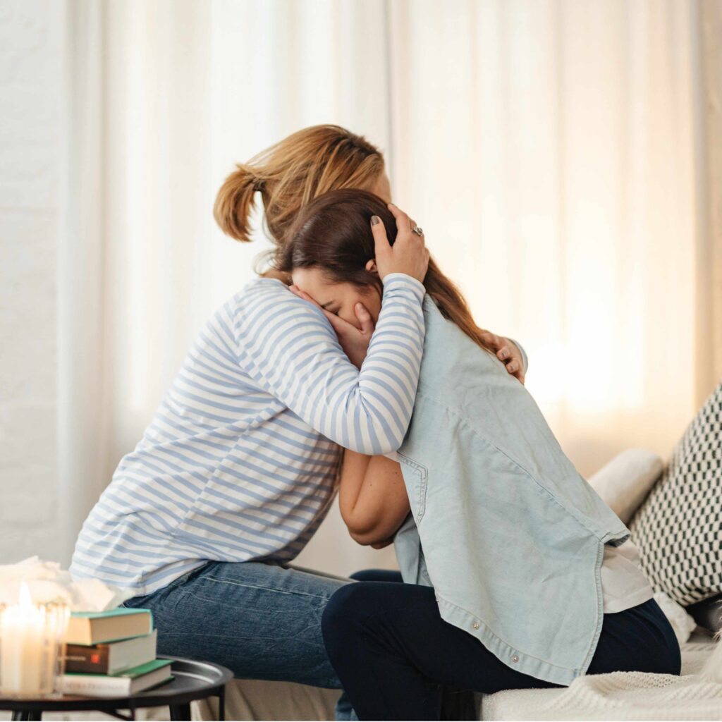 Auf dem Bild sind zwei Frau sehen. Die Frau auf der linken Seite nimmt die etwas jünger aussehende Frau, auf der rechten Seite, in den Arm. Die Frau auf der rechten Seite hält sich die Hände vor das Geschicht
